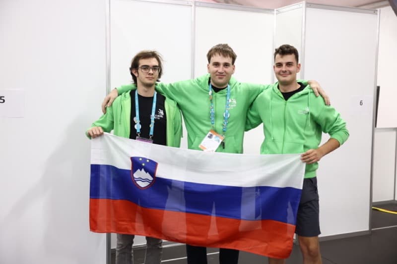 trije tekmovalci Vegove v zelenih puloverjih držijo slovensko zastavo