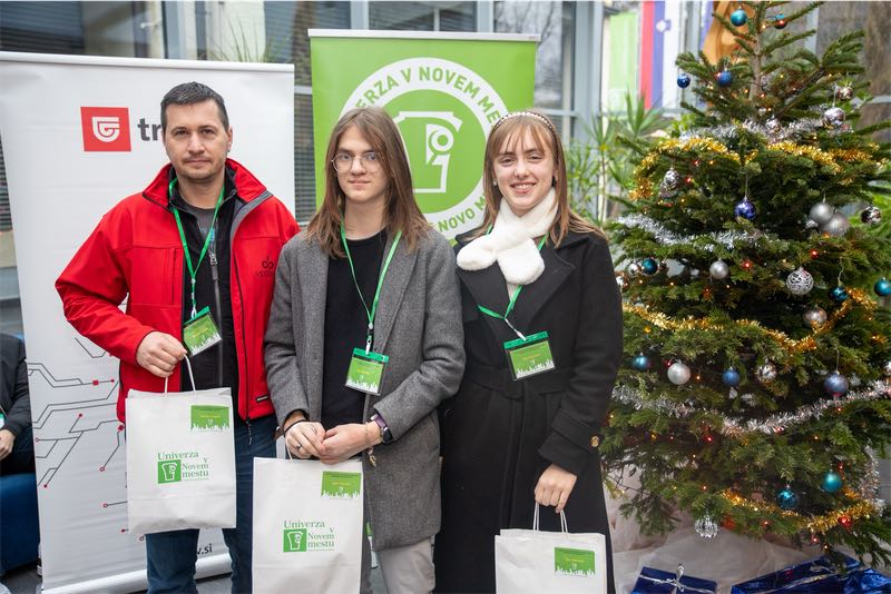 Tia Udovičić, Jure Maček in učitelj Marko Tomšič stojijo z darilnimi vrečkami pred okrašeno novoletno jelko