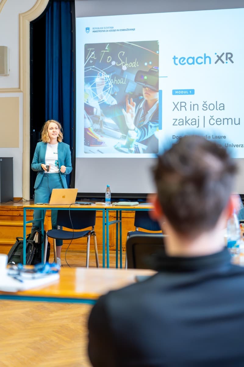 Predavateljica stoji pred platnom, na katerem piše XR v šoli, zakaj, čemu.