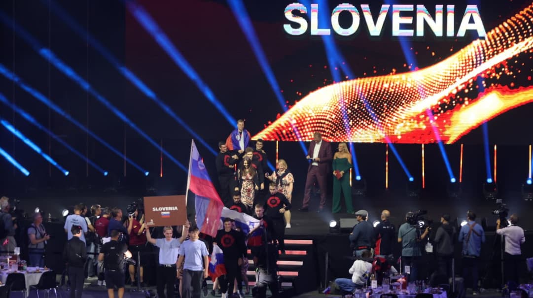 otvoritvena slovesnost. Na odru velika skupina tekmovalcev s svojimi zastavami. V ozadju pisani svetlobni efekti.