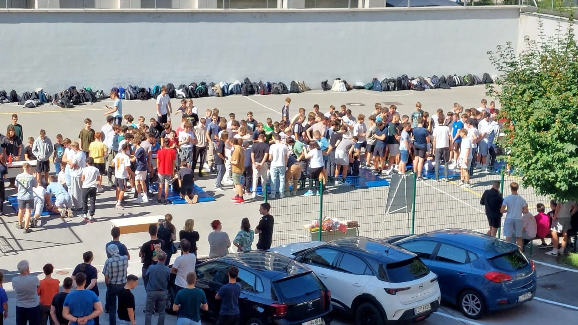 ob krstu prvošolcev na sončen dan, velika skupina dijakov stoji na šolskem igrišču. Za njimi lepo zloženi nahrbtniki ob steni igrišča. S strani jih opazuje jih skupina učiteljev.