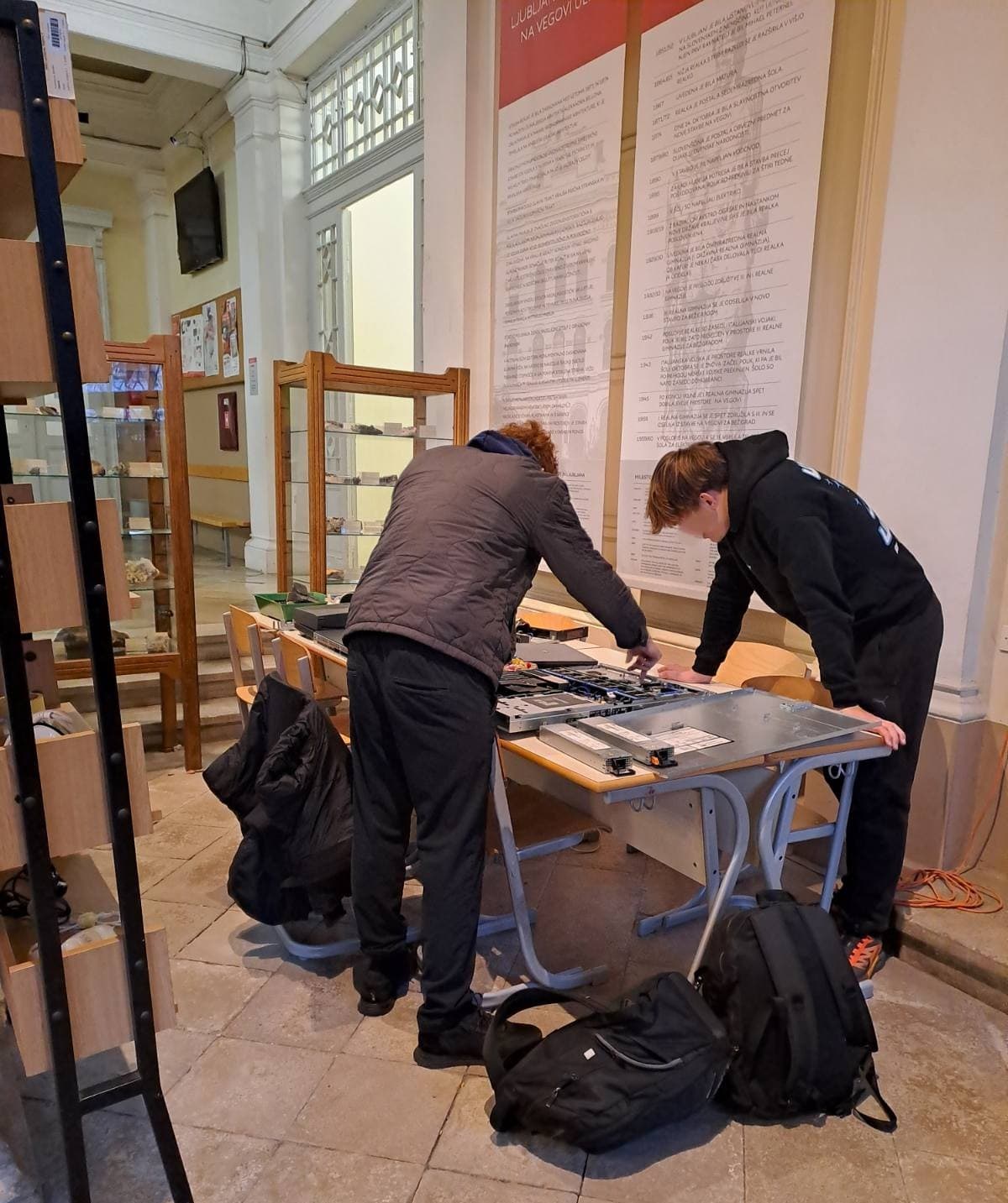 Popravljalnica v šolski avli, kjer dva dijaka pregledujeta strežnik. Na mizi je različno orodje za popravljanje elektronskih naprav. Na tleh sta nahrbtnika, levo regal s popravljenimi aparati in v ozadju minerali v steklenih vitrinah.