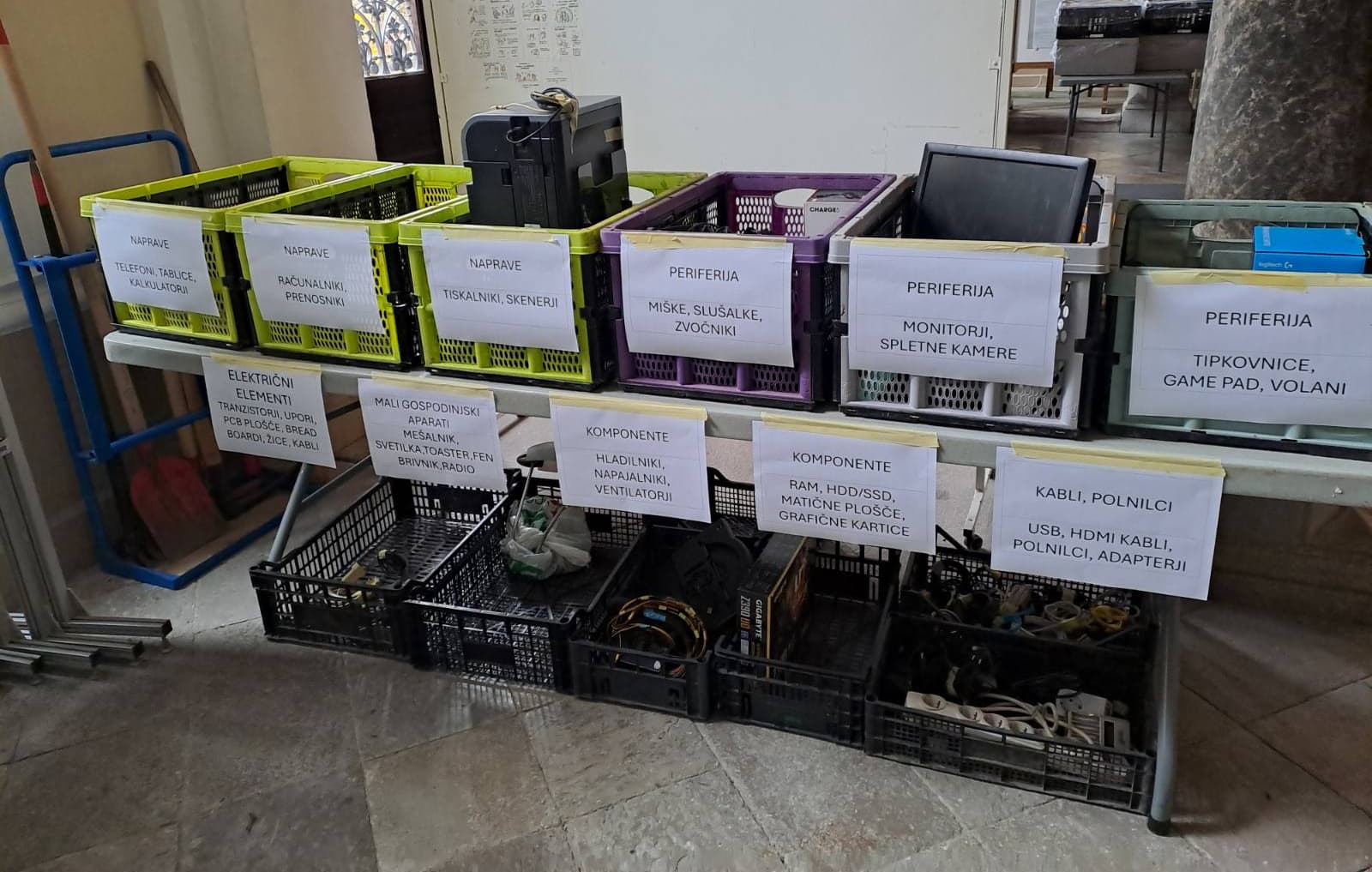 Zbirni center v šolski avli, kjer so na mizi z napisi označeni zaboji, v katerih so stara ali pokvarjena elektronska oprema (tiskalnik, monitor, zvočniki, razdelilci, kabli, napajalniki) in komponente. V ozadju so oglasna table in zaboja za šolsko malice, levo ob steni pa lopate za sneg.