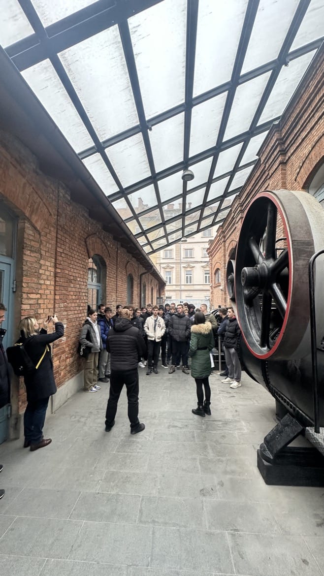 Skupina dijakov in dijakinj psluša razlago v dolgem prehodu med dvema zgodovinskima opečnatama stavbama z velikim okni.