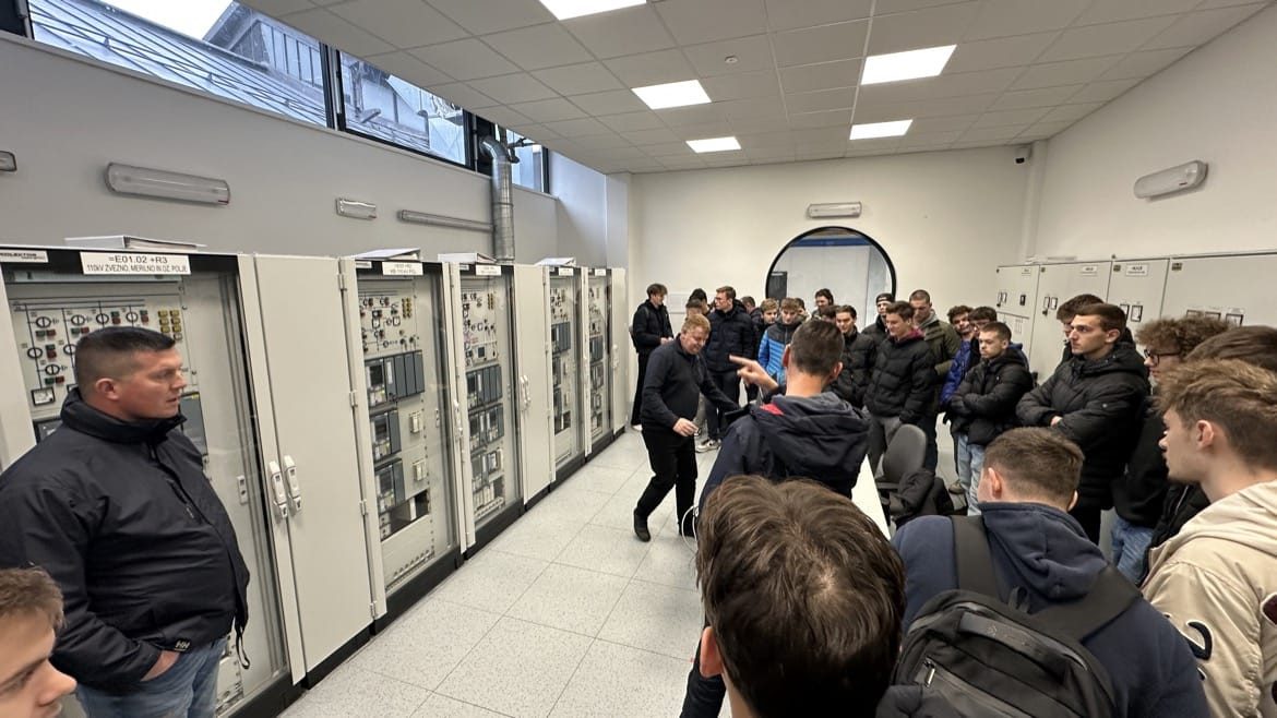 Večja skupina dijakov v nadzornem prostoru pred kontrolnimi ploščami z stikali in merilnimi inštrumenti. Poslušajo razlago zaposlenega upravljalca.