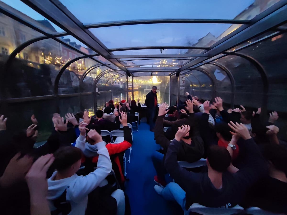 Dijaki na ladjici na plovbi po reki Ljubljanici, sedijo na stolih z dvignjenimi rokami zibaju v ritmu glasbe. V ozadju na obrežju z lučkami osvetljeno drevje in okolica.