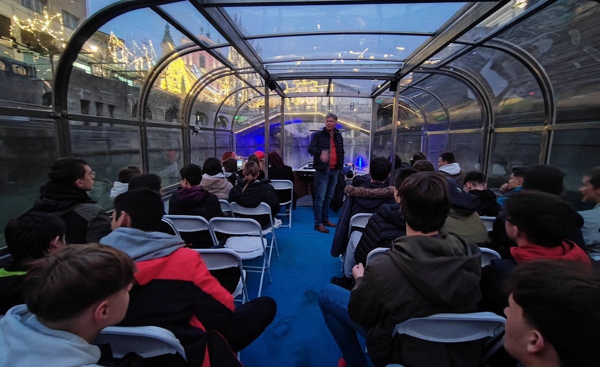 Dijaki sedijo na ladjici in opazujejo čarobno okrašeno Ljubljano. Vodnik nam razlaga zanimivosti. V ozadju na obrežju z lučkami osvetljeno drevje in okolica.