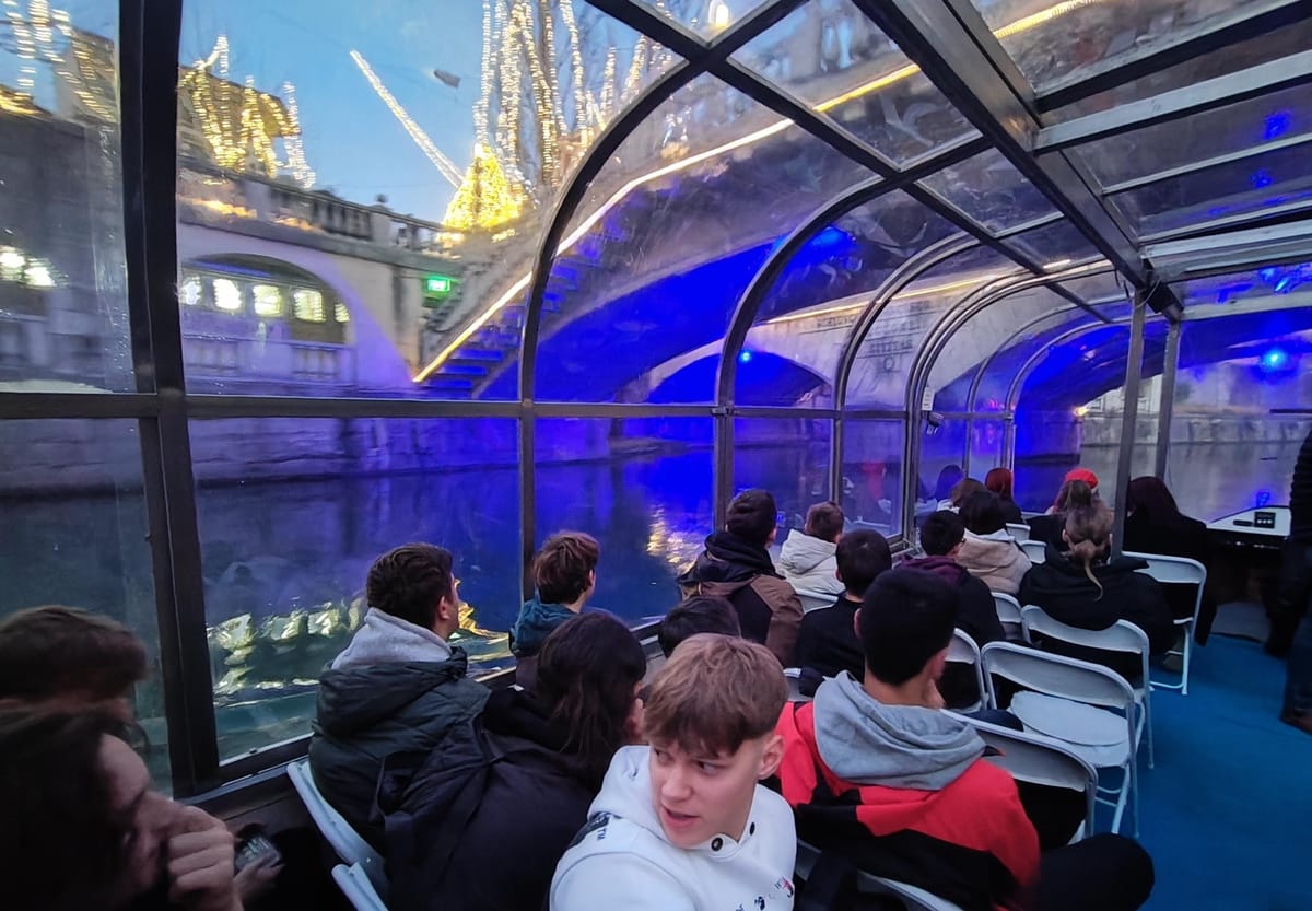 Dijaki sedijo na ladjici, plujejo pod tromostovjem. V ozadju na obrežju z lučkami osvetljeno drevje in okolica.