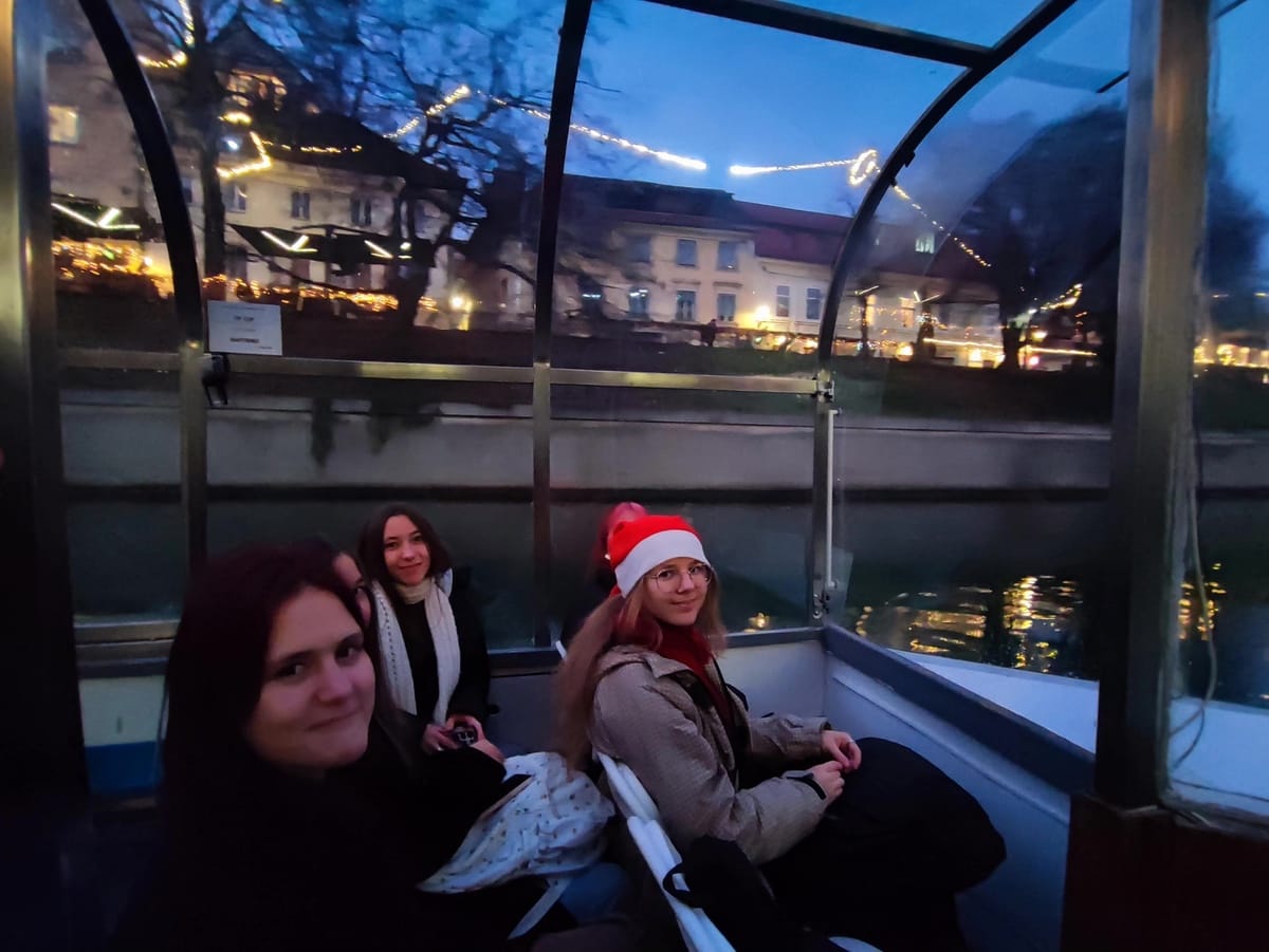 Tri dijakinje na vožnji z ladjico. Ena izmed njih nosi rdečo bočikovo kapo. V ozadju na obrežju z lučkami osvetljeno drevje in okolica.