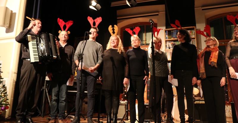 šolski pevski zbor devetih pevk in pevcev, svečano oblečenih, na zatemnjenem odru. Na glavi nosijo stilizirane jelenje rogove v duhu božičnih praznikov. Pevec na levi s harmoniko.