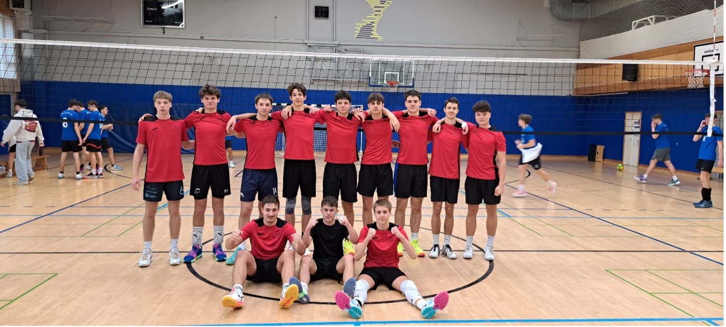 Skupina dvanajstih dijakov v dvorani pred odbojkarsko mrežo v rdeče črni športni opremi.