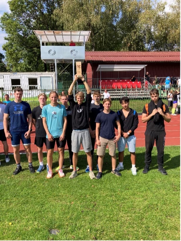 Skupinski portret devetih dijakov na zelenici, eden izmed njih drži dvignjen pokal. V ozadju tekaška steza.