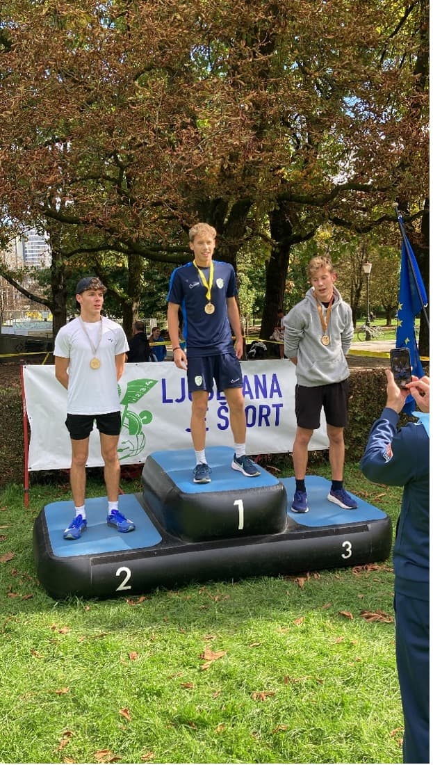 Trije dijaki na zelenici, v športni opremi na odru za zmagovalce. Okrog vratu imajo obešene prejete medalje.