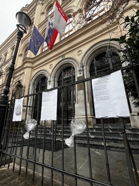 Pogled na glavni vhod šolske stavbe z obokanimi vrati. Nad vhodom visijo zastave, na ograji pred stopnicami pa je pritrjenih več listov z rokopisnimi besedili in risbami. Na ograji so ob rokopisih in karikaturah zavezani kozarci za vino.