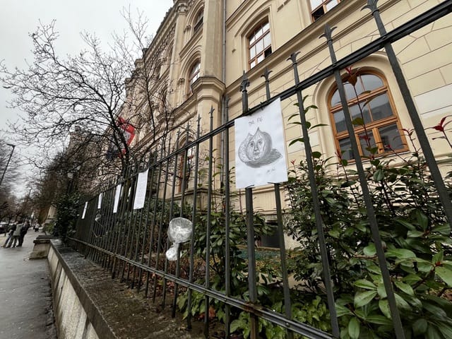 Daljši pogled ob ograji pred šolsko stavbo, ob kateri so pritrjeni listi z risbami. Na ograjo so obešeni vinski kozarci s portretom Franceta Prešerna. Ograjo spremlja zeleno grmičevje, ob pločniku pa se v ozadju vidi nekaj pešcev in gola drevesa v oblačnem vremenu.