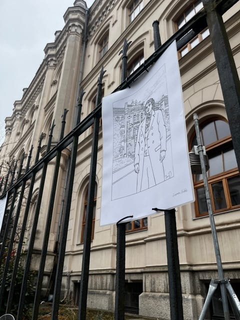 Na črni kovinski ograji pred zgodovinsko šolsko stavbo visi list papirja z črno belo risbo Franceta Prešerna v plašču, postavljenega pred fasado stavbe. Ograja je v ospredju, v ozadju pa so vidna velika okna in arhitekturni detajli stavbe.