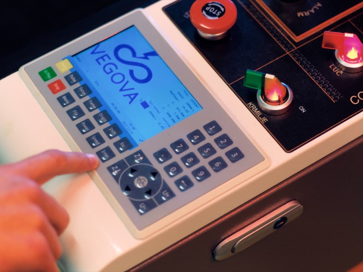 Bližnji posnetek roke, ki pritiska gumbe na industrijski nadzorni plošči z zaslonom na dotik na katerem je izrisan logotip Vegove in gumbom za izklop v sili.