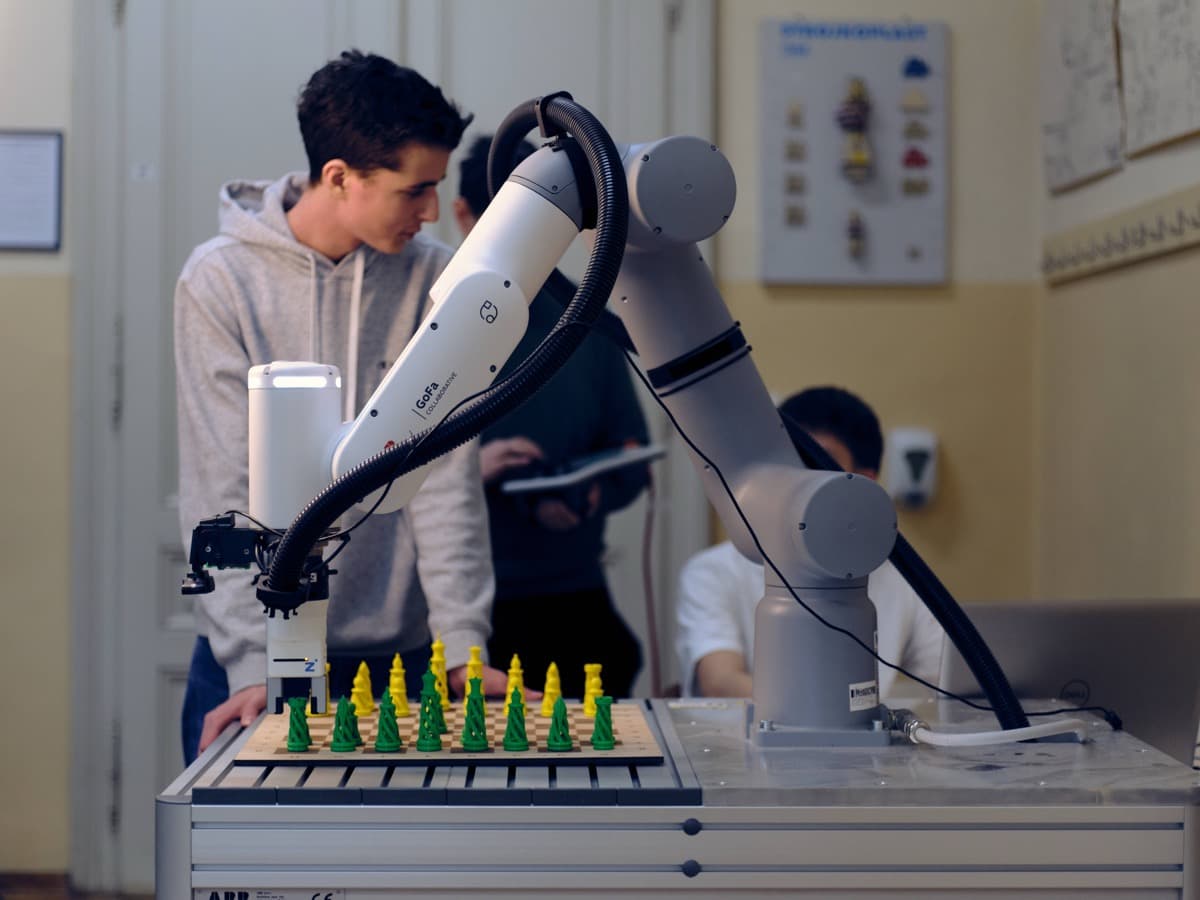 Dijak robotsko roko, postavljeno nad šahovnico z zelenimi in rumenimi figurami, pripravljenimi za igro.