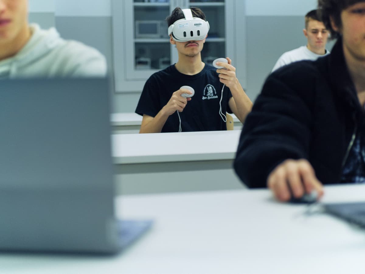 Dijak z očali za navidezno resničnost drži krmilnika in sedi za mizo v učilnici.