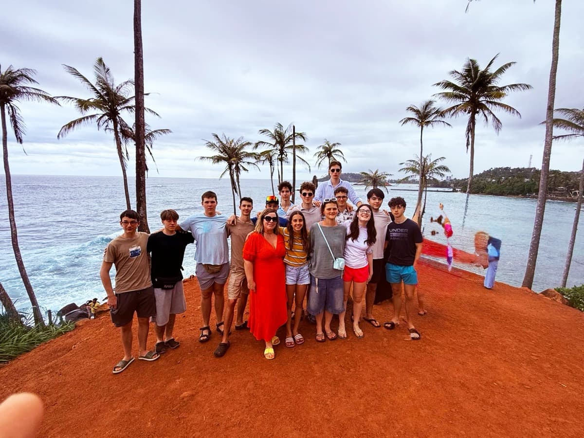 Skupina dijakov in profesorica stojijo na Coconut Beach ob kokosovih palmih in oceanu.