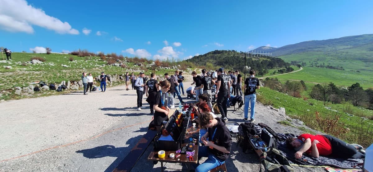 Skupina dijakov na prostem pripravlja in pregleduje tehnično opremo ob leseni mizi na makadamski poti v hriboviti pokrajini. V ozadju so zelena pobočja, drevesa in gore pod jasnim modrim nebom. Nekateri udeleženci stojijo in se pogovarjajo, drugi sedijo ali počivajo ob opremi.