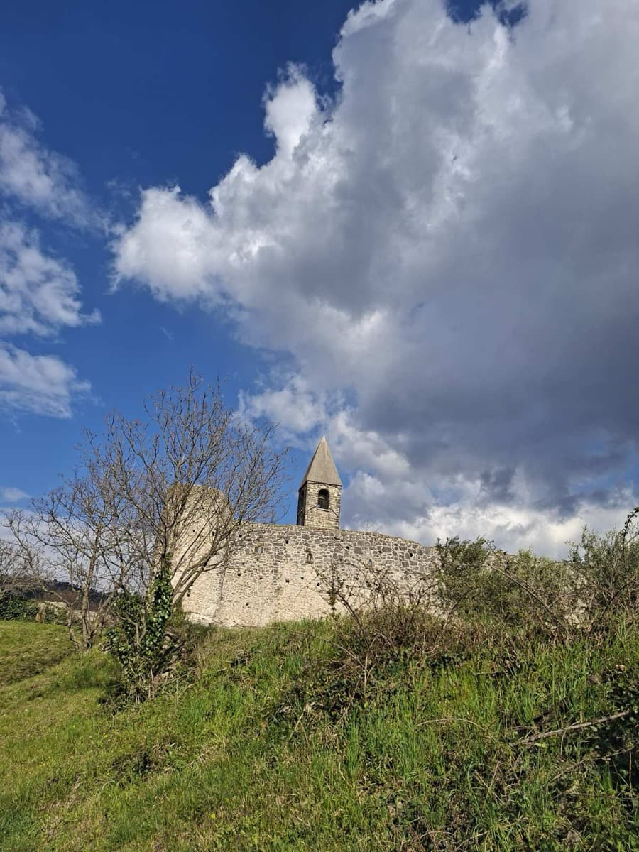 cerkveni zvonik in obrambni zid, nad njima modro nebo z belimi oblaki