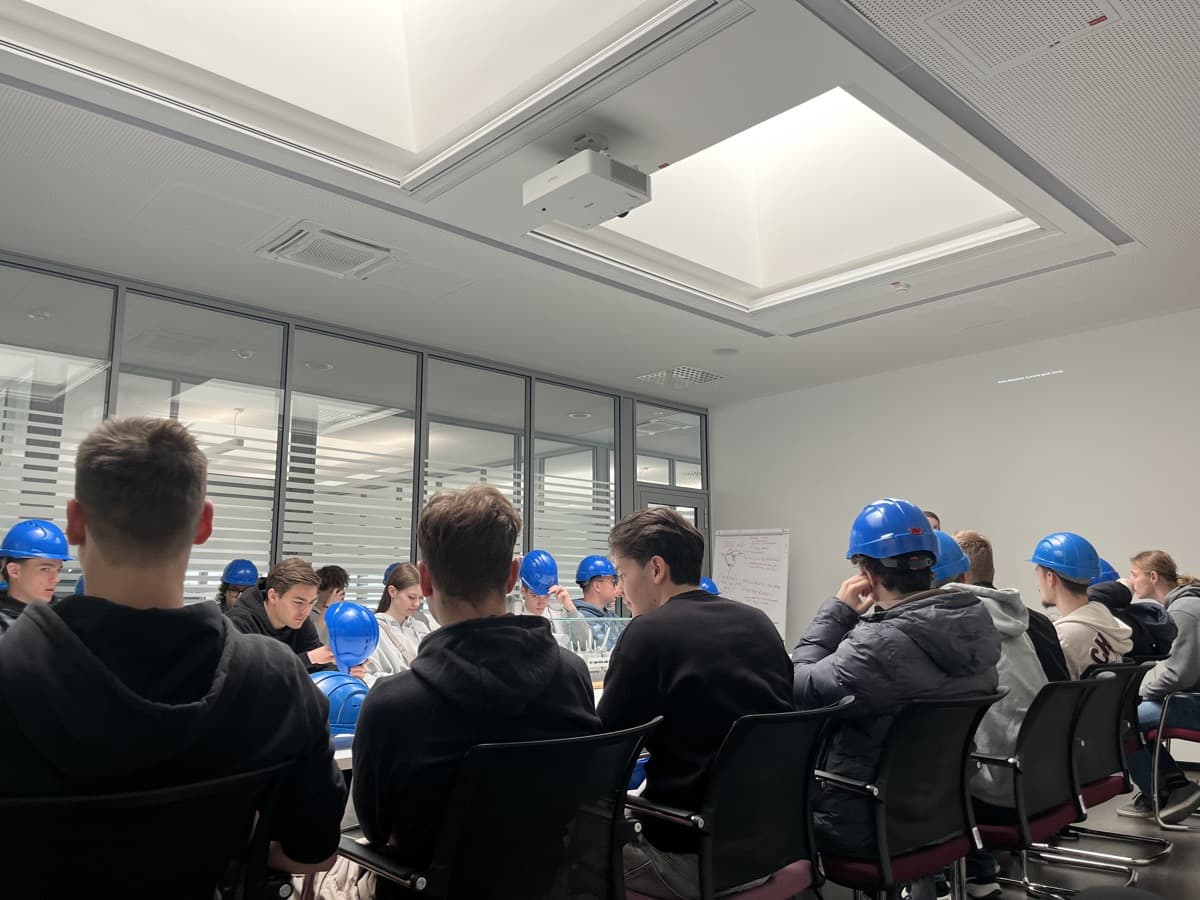 dijaki, ki sedijo v sejni sobi podjetja za mizo, nekateri imajo na glavah modre zaščitne čelade