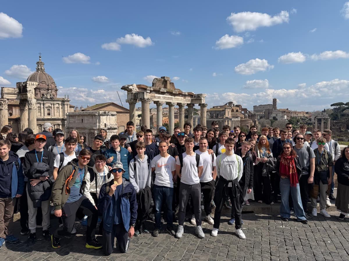 Skupina dijakov stoji na mestu arheoloških izkopavanj, za njimi so ostanki rimskega templja in drugih stavb s stebri. Na sončnem modrem nebu je nekaj belih oblačkov