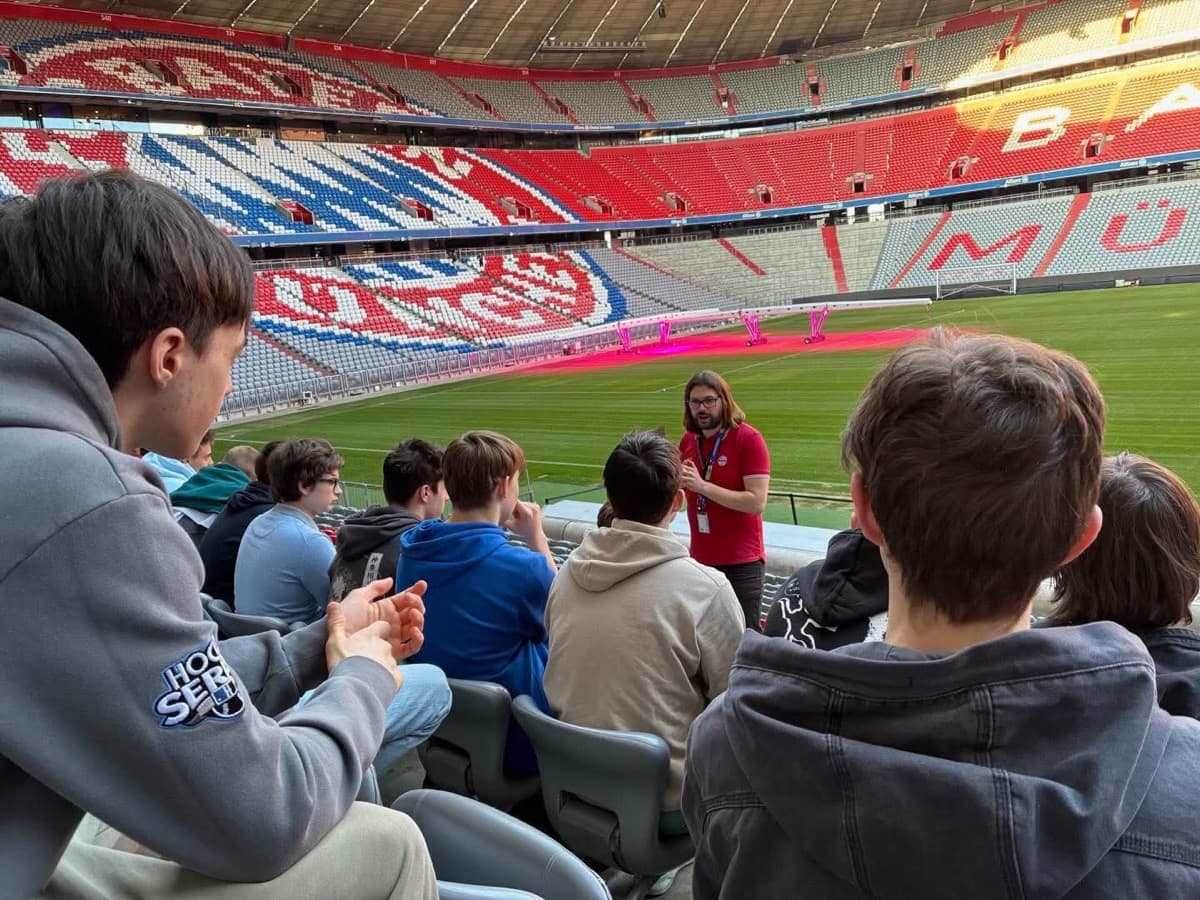 Sredi sedišča stadiona Allianz Arena stoji nemški vodnik v rdeči majici in z daljšimi lasmi, ki skupini sedečih dijakov razlaga o tem znamenitem stadionu. V ozadju sta nogometno igrišče z zeleno travo in nasprotni del praznega avditorija, obarvan z velikim logotipom Bayerna v rdeče-belo-modrih barvah