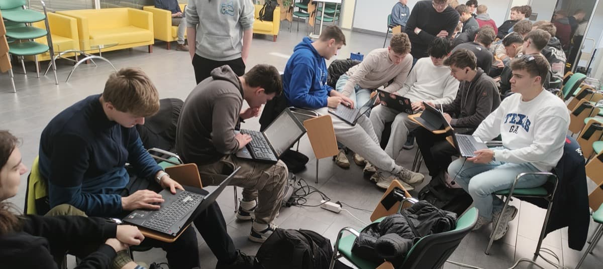 skupina dijakov, ki sedijo v polkrogu; večina ima na kolenih prenosne računalnike; tipkajo ali opazujejo ekran. Za njimi je še ena taka skupina. Prostor je podoben predavalnici, v ozadju so rumeni kavči in zeleni stoli, naloženi drug na drugega